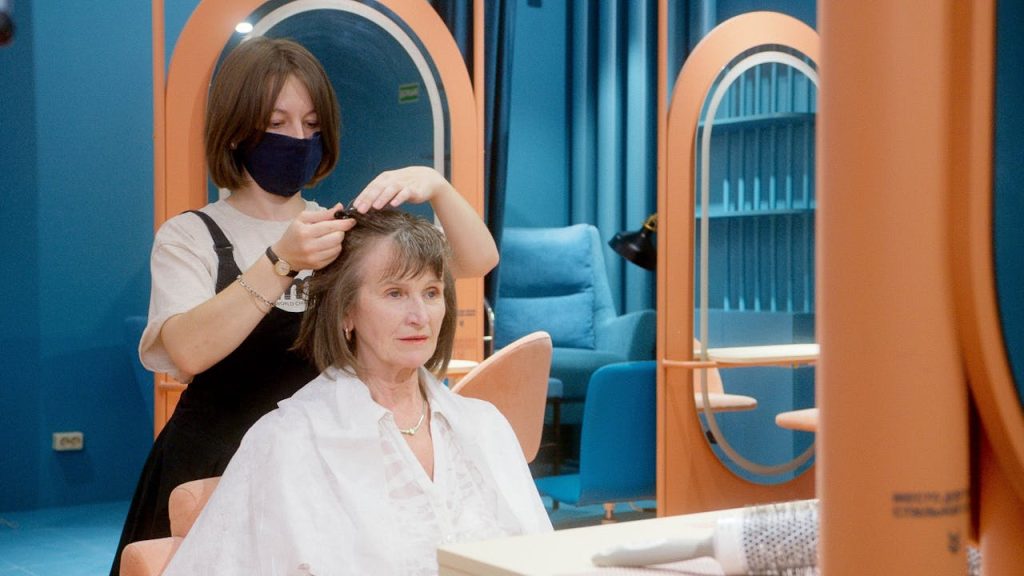 A senior woman receives styling at a modern hair salon by a masked hairstylist.